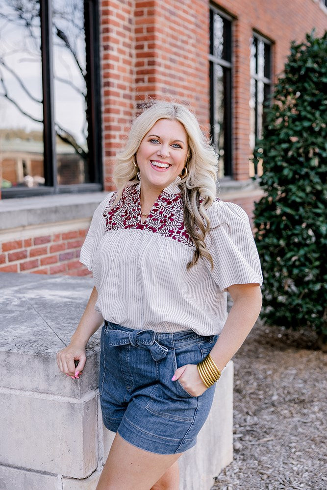 Woman with blonde hair standing in a front profile position wearing the Reece Top on campus. The focal point of this image is the embroidery on chest and lighting. The model paired this top with dark-wash denim shorts. 