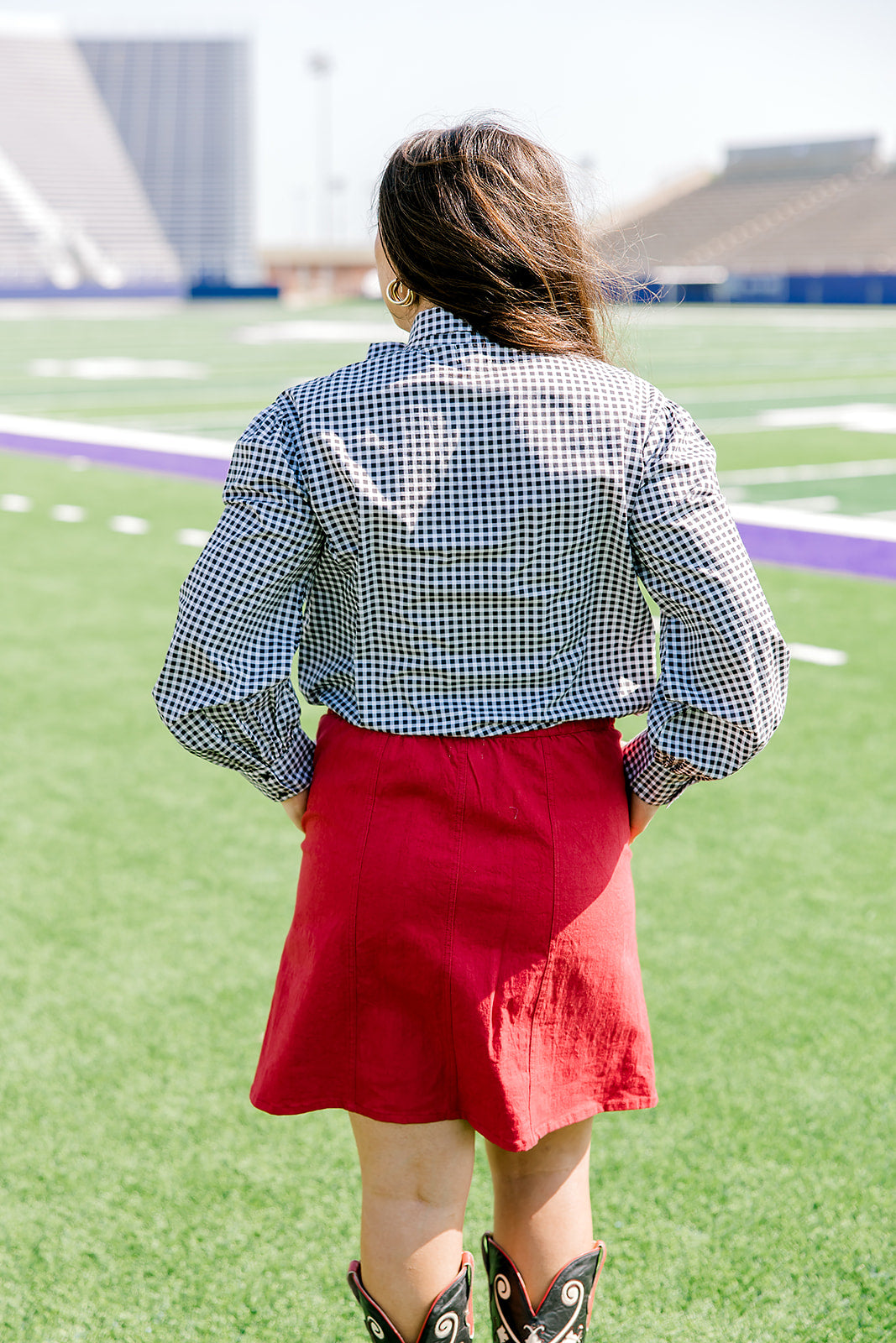 Woman with dark hair standing in a back profile position wearing the Sadie Top in a lifestyle scene. The focal point of this image is the black gingham pattern on back of top. The model paired this top with a red skirt.