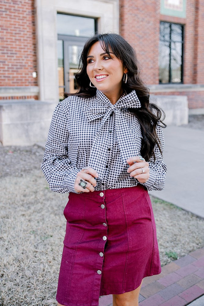 Woman with dark hair standing in a front profile position wearing the Sadie Top in a lifestyle scene. The focal point of this image is the black gingham pattern and bow that's tied on front of top. The model paired this top with a maroon skirt. 