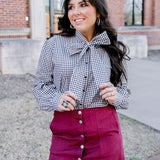 Woman with dark hair standing in a front profile position wearing the Sadie Top in a lifestyle scene. The focal point of this image is the black gingham pattern and bow that's tied on front of top. The model paired this top with a maroon skirt. 