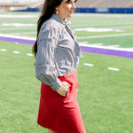Woman with dark hair standing in a side profile position wearing the Sadie Top in a lifestyle scene. The focal point of this image is the black gingham pattern as well the bow on front of top. The model paired this top with a red skirt.