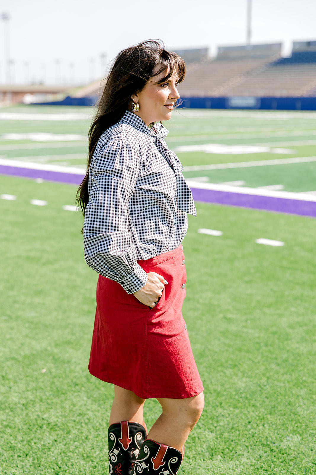 Woman with dark hair standing in a side profile position wearing the Sadie Top in a lifestyle scene. The focal point of this image is the black gingham pattern as well the bow on front of top. The model paired this top with a red skirt.
