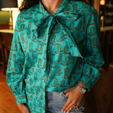 Woman with dark hair wearing the Sadie Body style with brown boots graphics along top and bow. Model paired this top with light-wash denim jeans. 