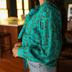 Woman with dark hair wearing the Sadie Body style with brown boots graphics along top and bow. Model paired this top with light-wash denim jeans. 