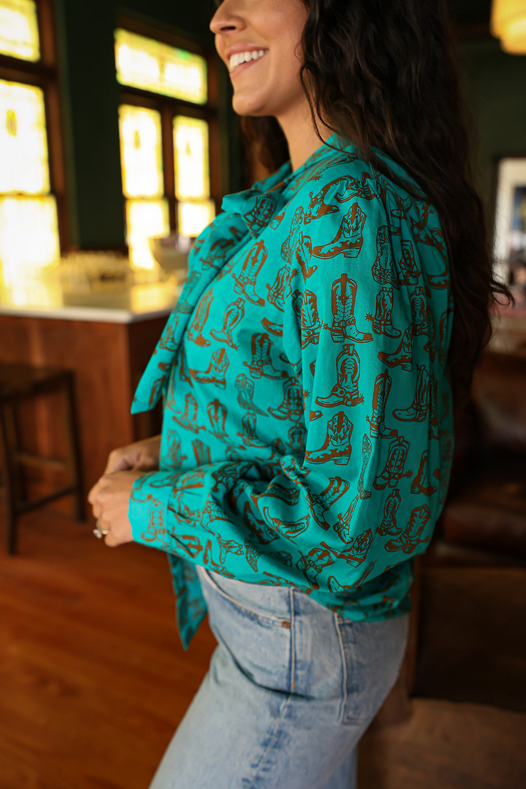 Woman with dark hair wearing the Sadie Body style with brown boots graphics along top and bow. Model paired this top with light-wash denim jeans. 