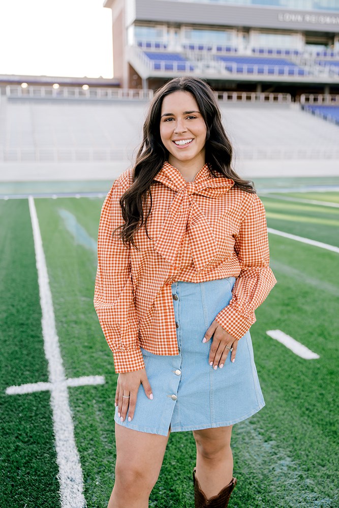 Woman with dark hair wearing a burnt orange gingham long-sleeve top, and light-wash denim shorts. 