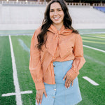 Woman with dark hair wearing a burnt orange gingham long-sleeve top, and light-wash denim shorts. 