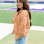 Woman with dark hair wearing a burnt orange gingham long-sleeve top, and light-wash denim shorts. 