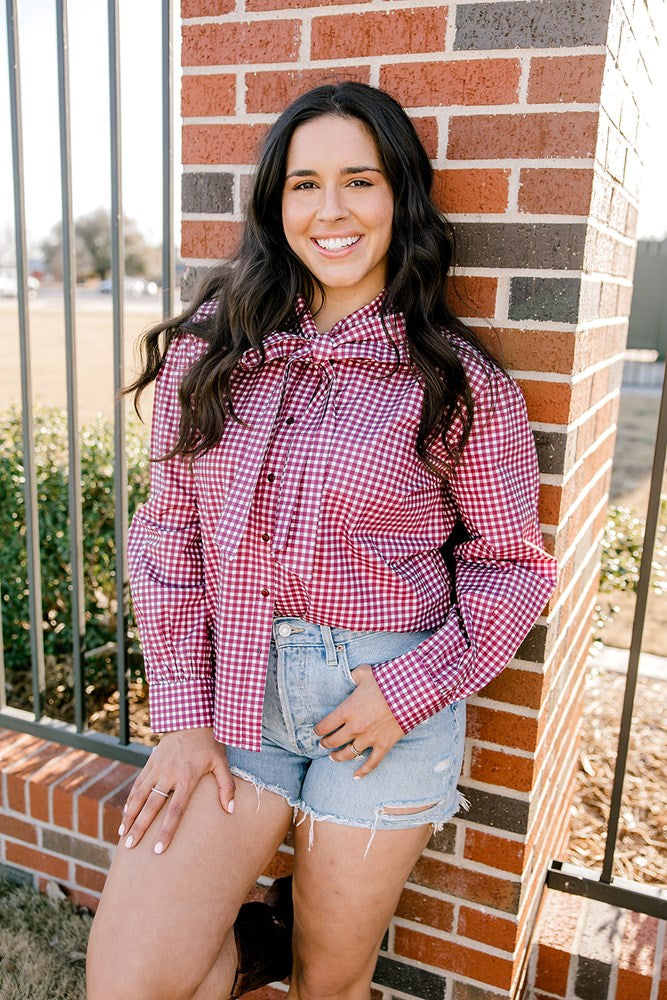 Woman with dark hair wearing a button down, maroon gingham long-sleeve with a bow tied in front, and light-wash denim shorts. 