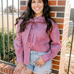 Woman with dark hair wearing a button down, maroon gingham long-sleeve with a bow tied in front, and light-wash denim shorts. 
