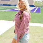 Woman with blonde hair standing in a side profile position wearing the Sadie Top in a lifestyle scene. The focal point of this image is the maroon gingham pattern along shirt, cuffed sleeve, and bow tied in front. The model paired this top with light-wash denim shorts. 