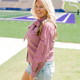 Woman with blonde hair standing in a side profile position wearing the Sadie Top in a lifestyle scene. The focal point of this image is the maroon gingham pattern along shirt, cuffed sleeve, and bow tied in front. The model paired this top with light-wash denim shorts. 