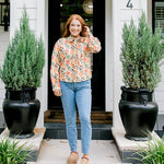 Woman with red hair wearing a long-sleeve top with multi-color ornament graphics & a bow-tied in the front. Model paired this top with light-wash denim jeans. 