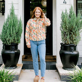 Woman with red hair wearing a long-sleeve top with multi-color ornament graphics & a bow-tied in the front. Model paired this top with light-wash denim jeans. 