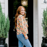 Woman with red hair wearing a long-sleeve top with multi-color ornament graphics & a bow-tied in the front. Model paired this top with light-wash denim jeans. 