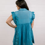 Woman wearing a blue dress with ruffled sleeves against a white background facing back. 
