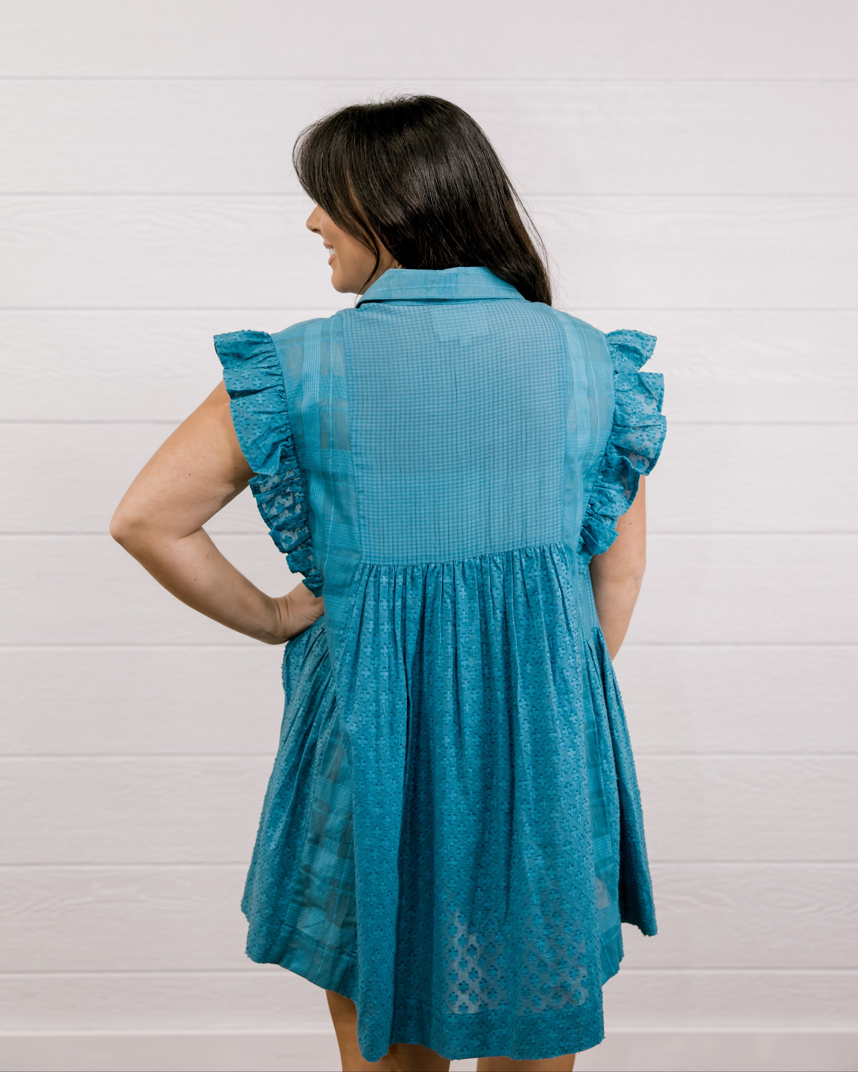Woman wearing a blue dress with ruffled sleeves against a white background facing back. 