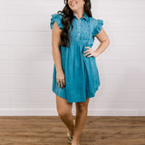 Woman wearing a blue dress standing on a wooden floor with a white wall background facing forward.