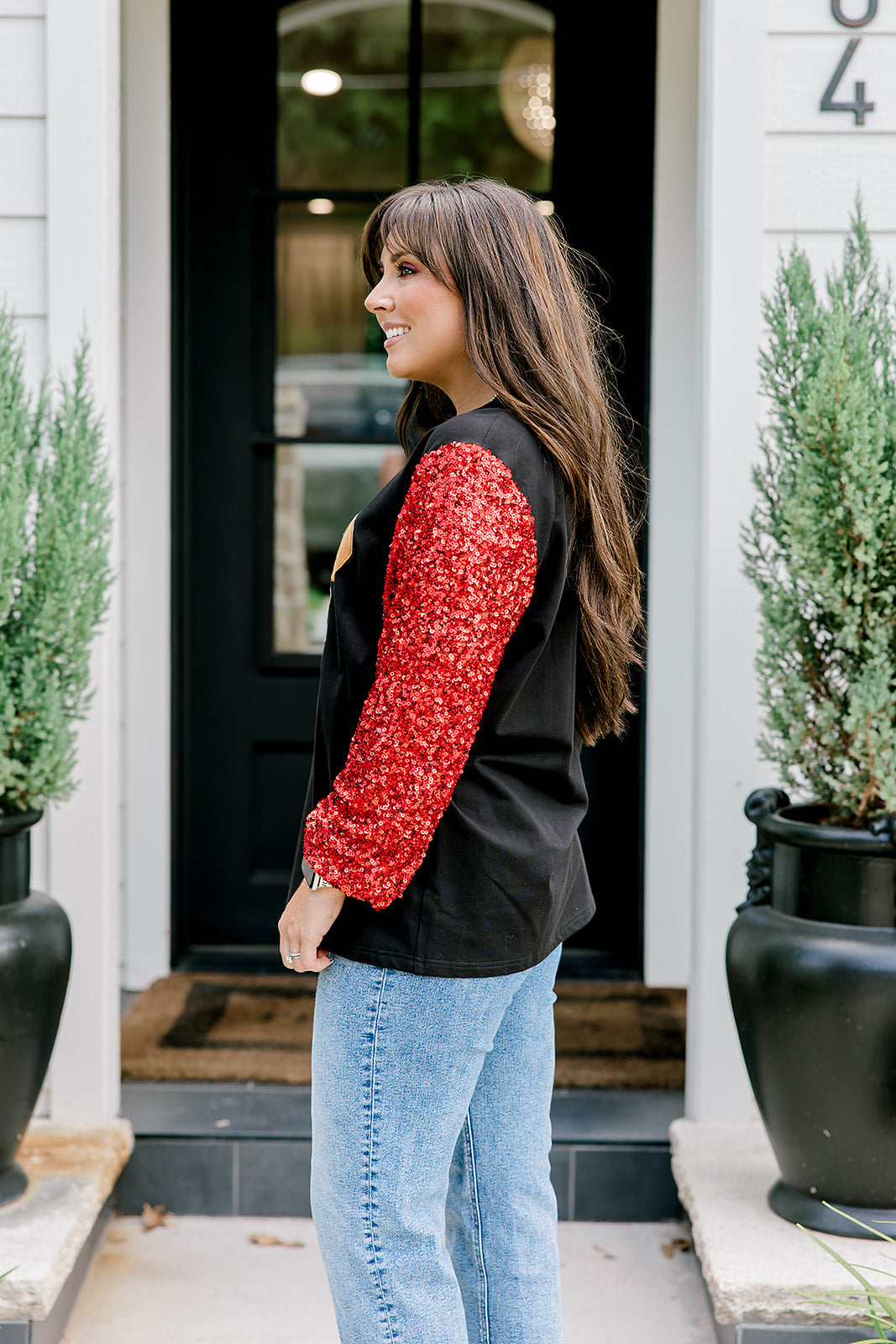 Woman with dark hair wearing long-sleeve top with embroidered cowboy Santa on front and red sparkle sleeves. Model paired this top with light-wash denim jeans. 