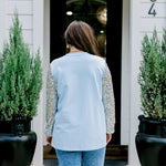 Woman with dark hair wearing a baby blue sweatshirt with iridescent sparkle sleeves; front of top has holiday truck embroidered on it. Model paired this top with light-wash denim jeans. 