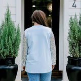 Woman with dark hair wearing a baby blue sweatshirt with iridescent sparkle sleeves; front of top has holiday truck embroidered on it. Model paired this top with light-wash denim jeans. 