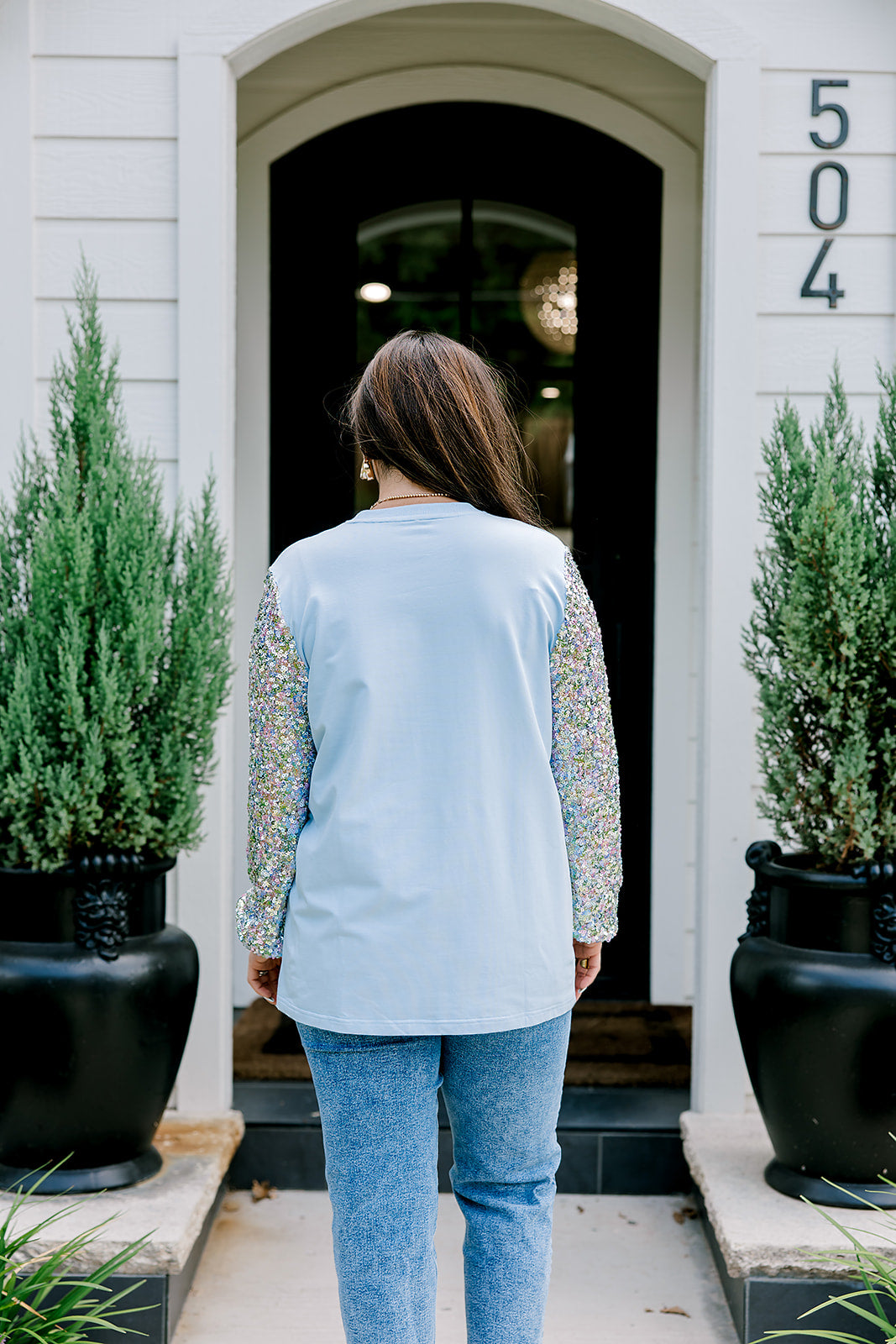 Woman with dark hair wearing a baby blue sweatshirt with iridescent sparkle sleeves; front of top has holiday truck embroidered on it. Model paired this top with light-wash denim jeans. 