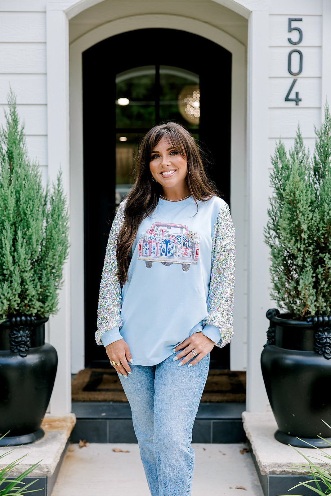 Woman with dark hair wearing a baby blue sweatshirt with iridescent sparkle sleeves; front of top has holiday truck embroidered on it. Model paired this top with light-wash denim jeans. 