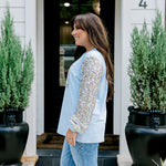 Woman with dark hair wearing a baby blue sweatshirt with iridescent sparkle sleeves; front of top has holiday truck embroidered on it. Model paired this top with light-wash denim jeans. 