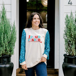 Woman with dark hair wearing a sweatshirt with turquoise sparkle sleeves & embroidery on front that says "Jesus Is The Reason". Model paired this top with dark-wash denim jeans. 