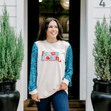 Woman with dark hair wearing a sweatshirt with turquoise sparkle sleeves & embroidery on front that says "Jesus Is The Reason". Model paired this top with dark-wash denim jeans. 