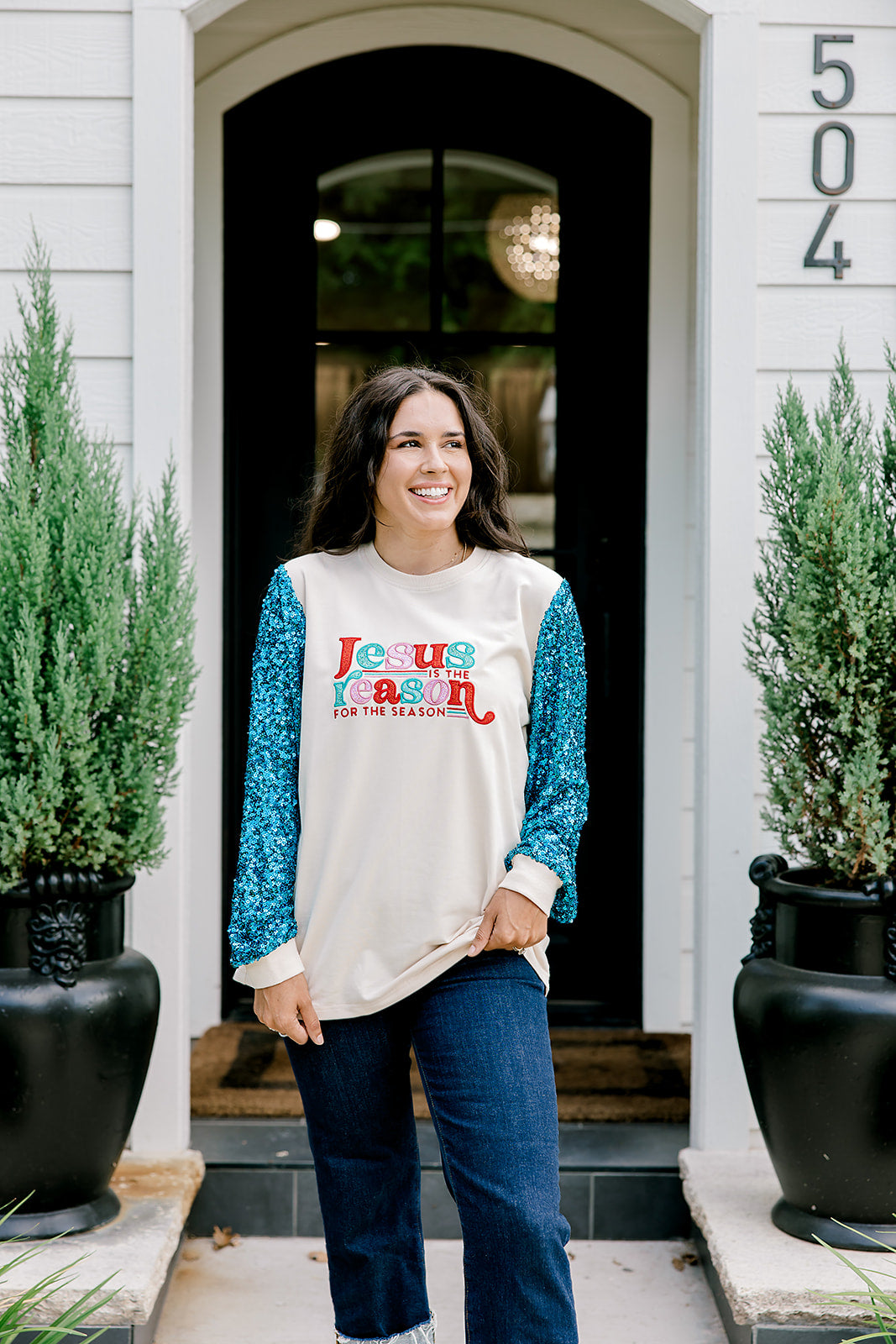 Woman with dark hair wearing a sweatshirt with turquoise sparkle sleeves & embroidery on front that says "Jesus Is The Reason". Model paired this top with dark-wash denim jeans. 