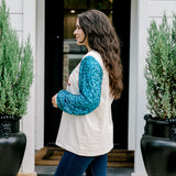 Woman with dark hair wearing a sweatshirt with turquoise sparkle sleeves & embroidery on front that says "Jesus Is The Reason". Model paired this top with dark-wash denim jeans. 