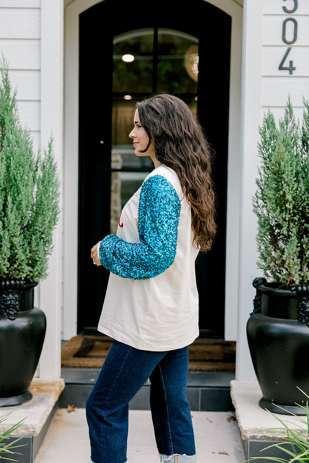 Woman with dark hair wearing a sweatshirt with turquoise sparkle sleeves & embroidery on front that says "Jesus Is The Reason". Model paired this top with dark-wash denim jeans. 