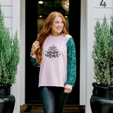 Woman with red hair wearing a sweater with green sparkle sleeves, and multi-color tree embroidery on front with words "Merry & Bright" on front. Model paired this top with faux leather leggings. 
