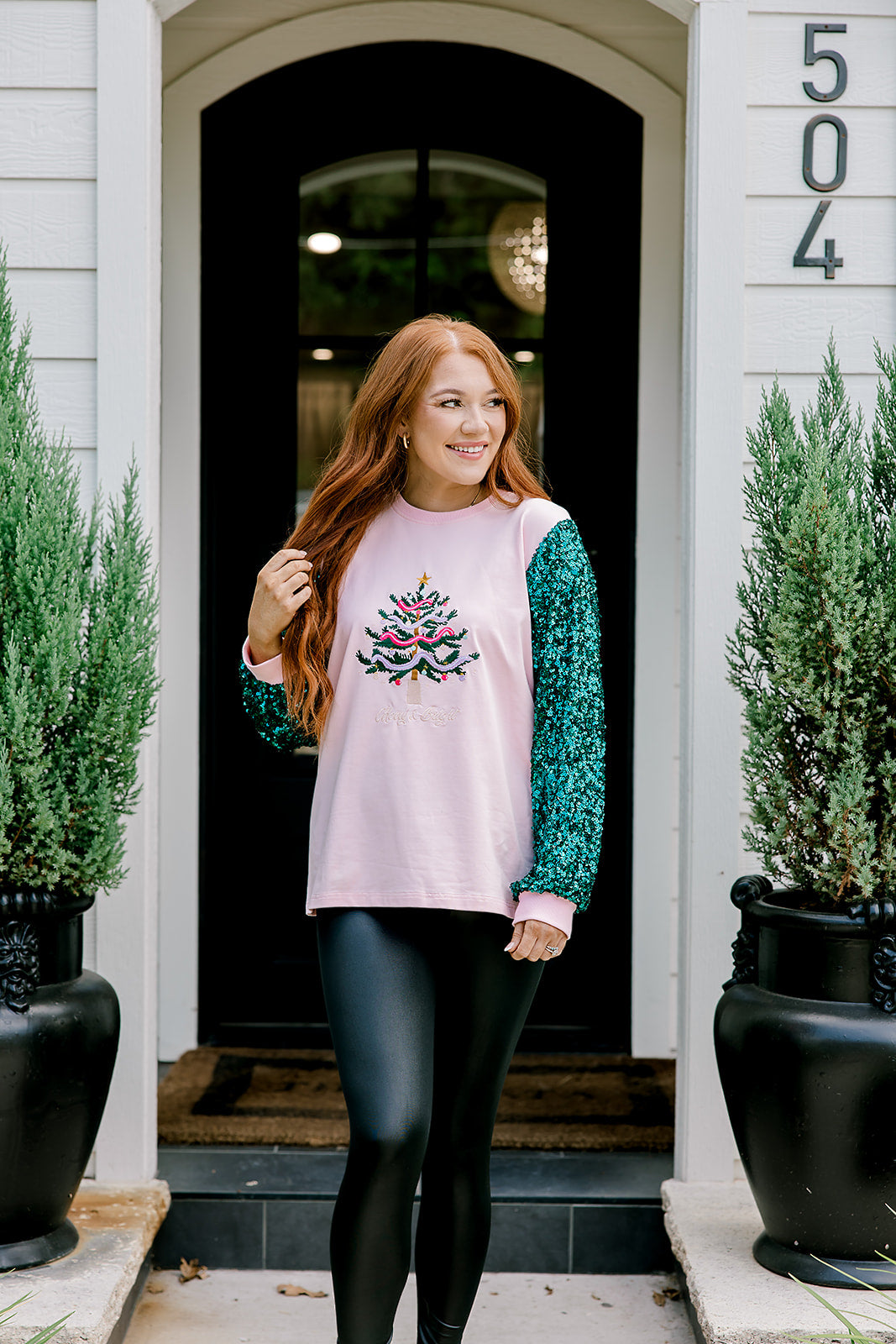 Woman with red hair wearing a sweater with green sparkle sleeves, and multi-color tree embroidery on front with words "Merry & Bright" on front. Model paired this top with faux leather leggings. 