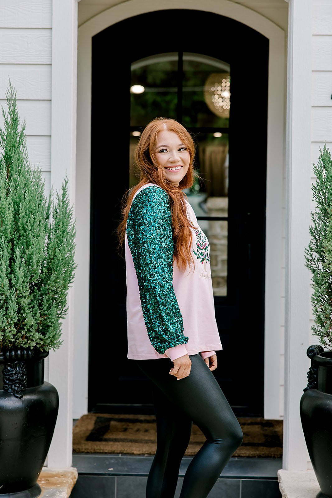 Woman with red hair wearing a sweater with green sparkle sleeves, and multi-color tree embroidery on front with words "Merry & Bright" on front. Model paired this top with faux leather leggings. 