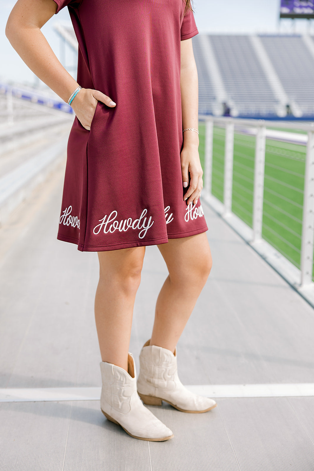 Woman wearing a midi maroon dress with howdy embroidered in white. The model paired this dress with cream boots. 
