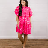 Woman wearing a pink polka dot dress standing against a white wall facing the front. 