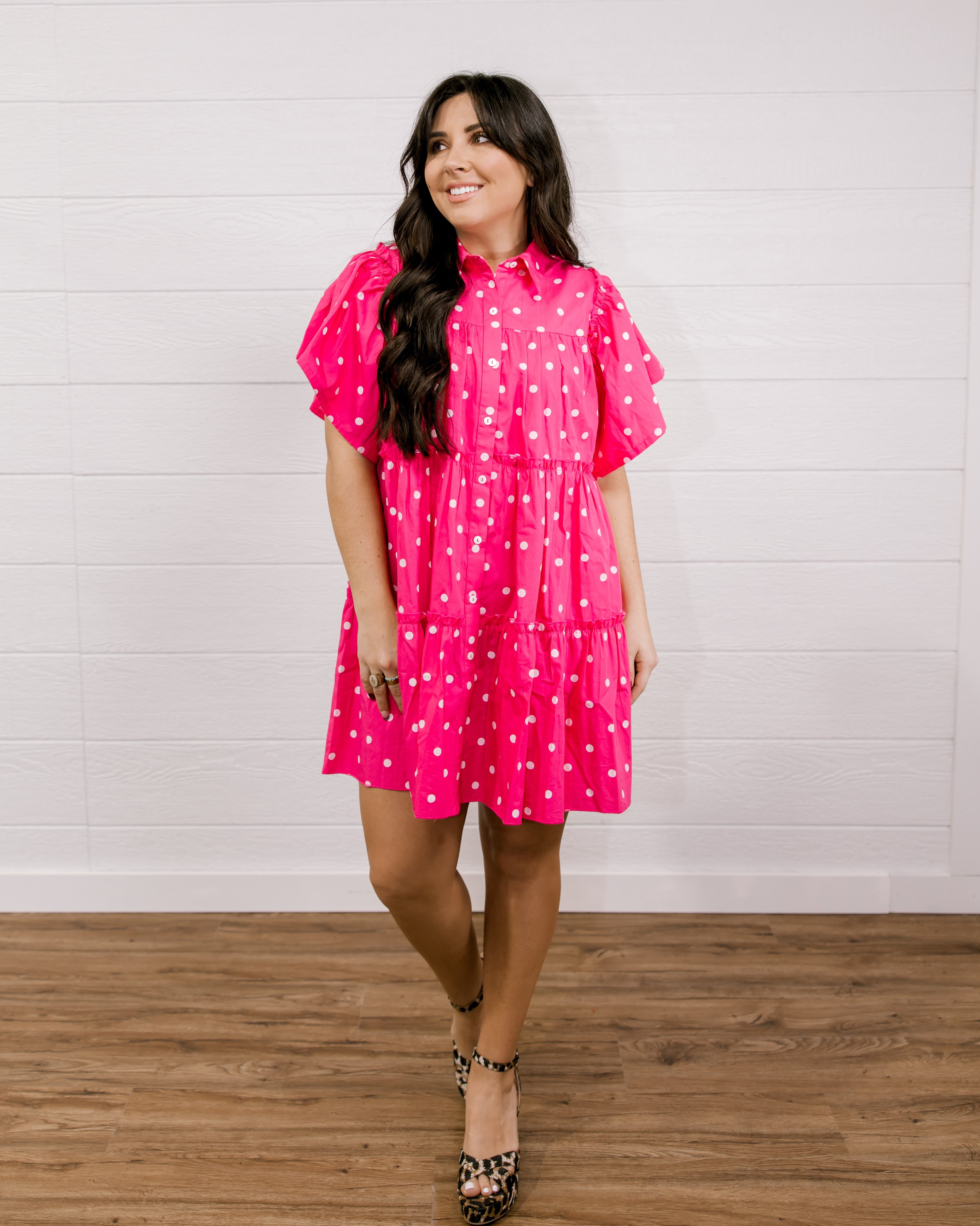 Woman wearing a pink polka dot dress standing against a white wall facing the front. 