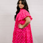 Woman wearing a pink dress with white polka dots against a light gray background facing side. 
