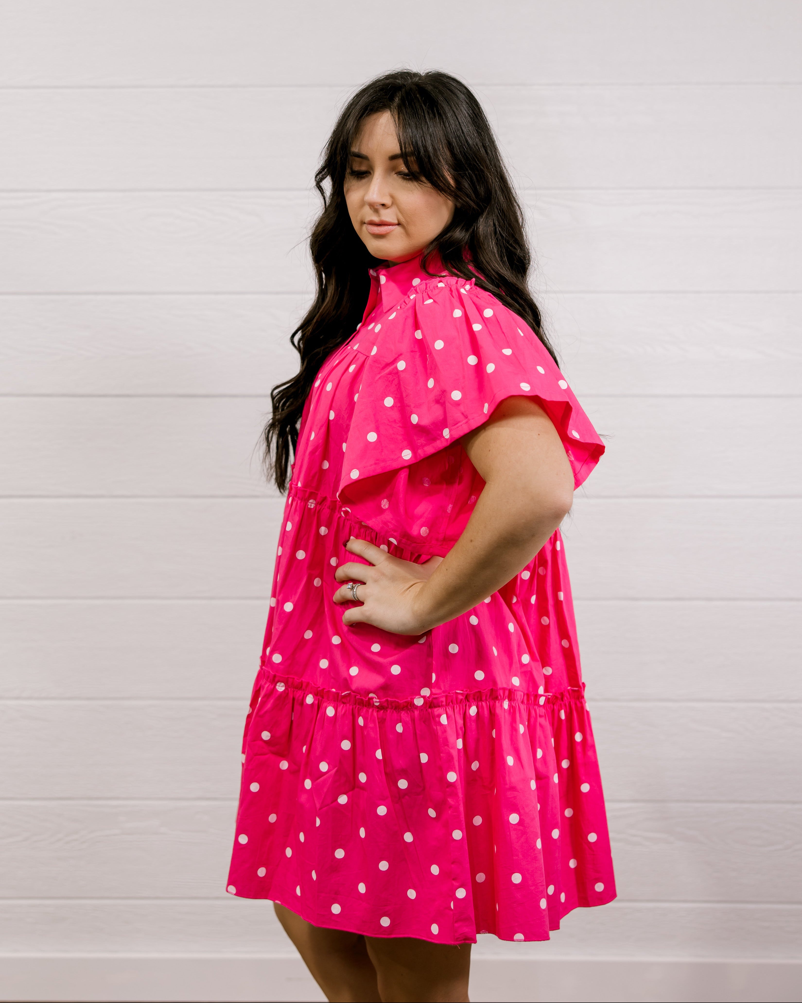 Woman wearing a pink dress with white polka dots against a light gray background facing side. 