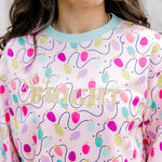 Woman with dark hair wearing a sweatshirt with Christmas light graphics  that are multi-colored. Model paired this top with dark-wash denim jeans. 