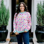 Woman with dark hair wearing a sweatshirt with Christmas light graphics  that are multi-colored. Model paired this top with dark-wash denim jeans. 