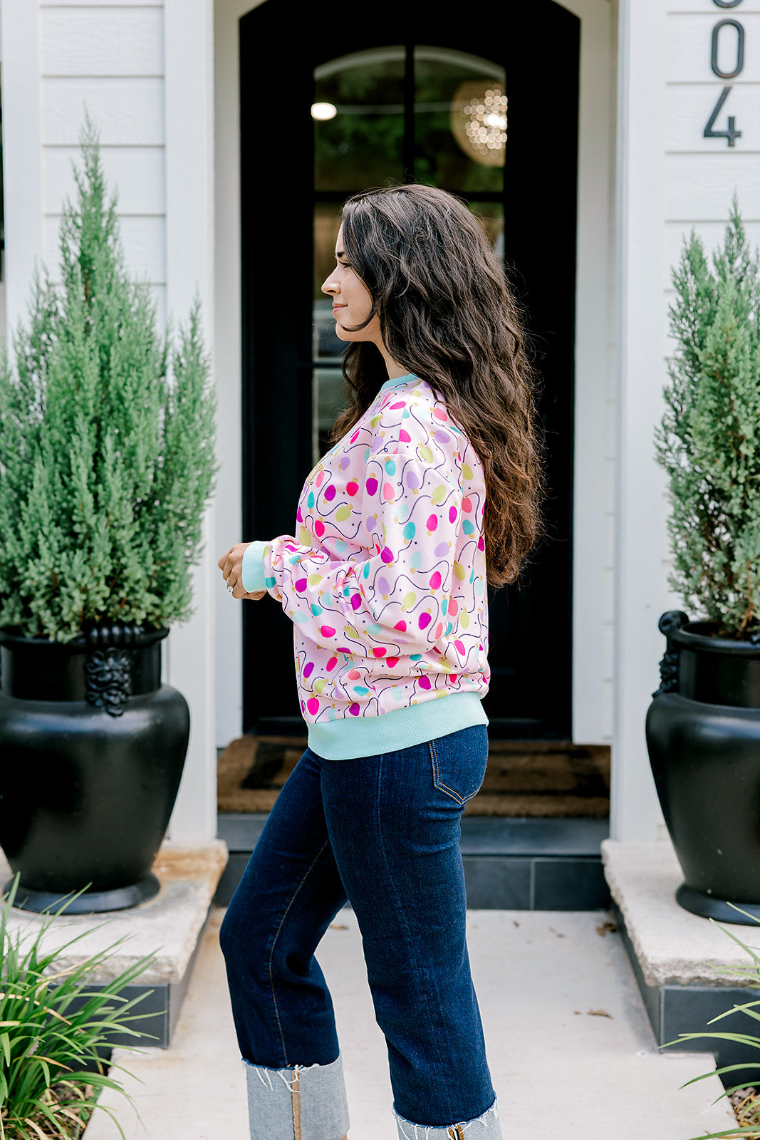 Woman with dark hair wearing a sweatshirt with Christmas light graphics  that are multi-colored. Model paired this top with dark-wash denim jeans. 