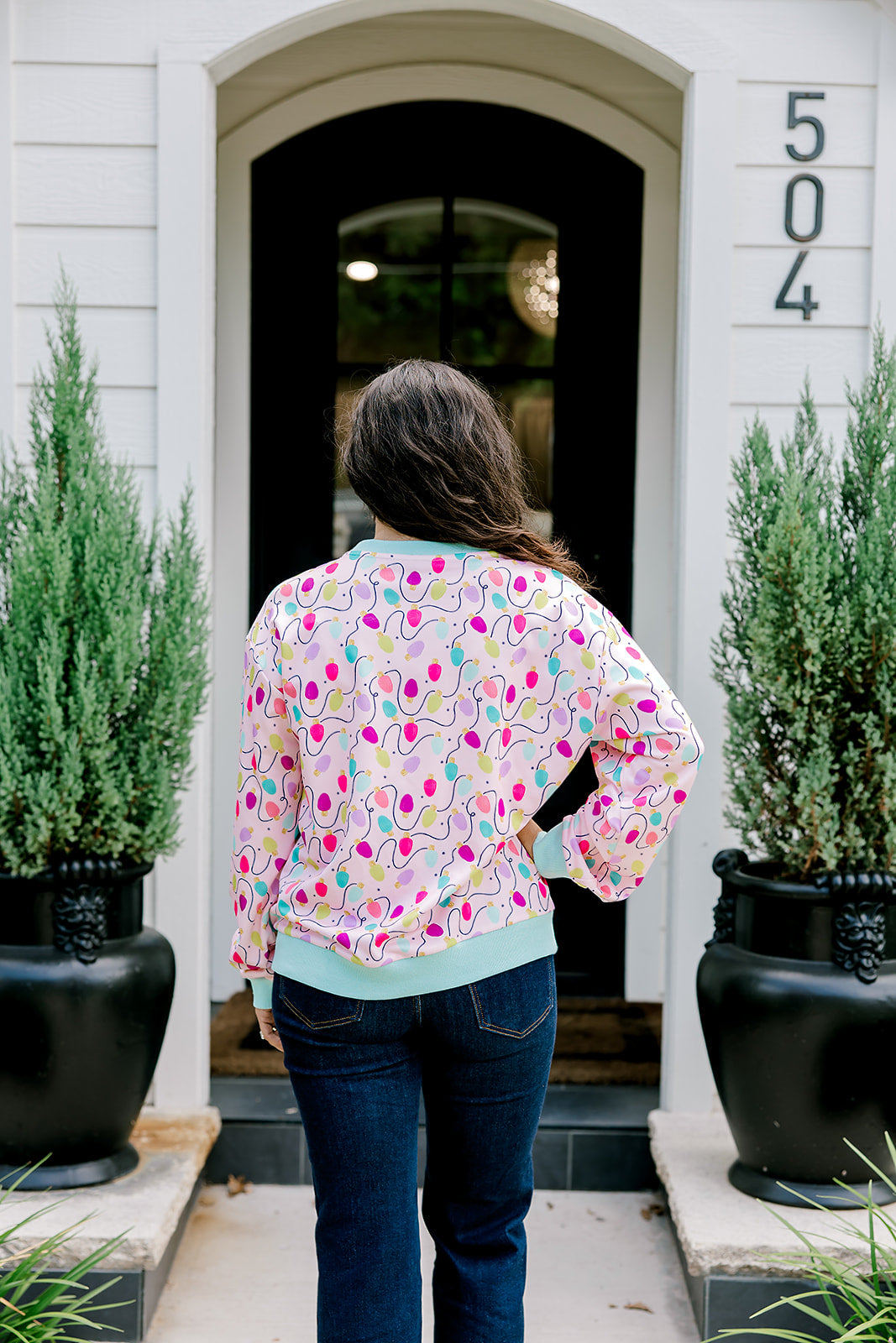 Woman with dark hair wearing a sweatshirt with Christmas light graphics  that are multi-colored. Model paired this top with dark-wash denim jeans. 