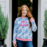 Woman with red hair wearing a blue floral sweatshirt with pink accents and "Merry Christmas" embroidered on front. Model paired this top with denim jeans. 