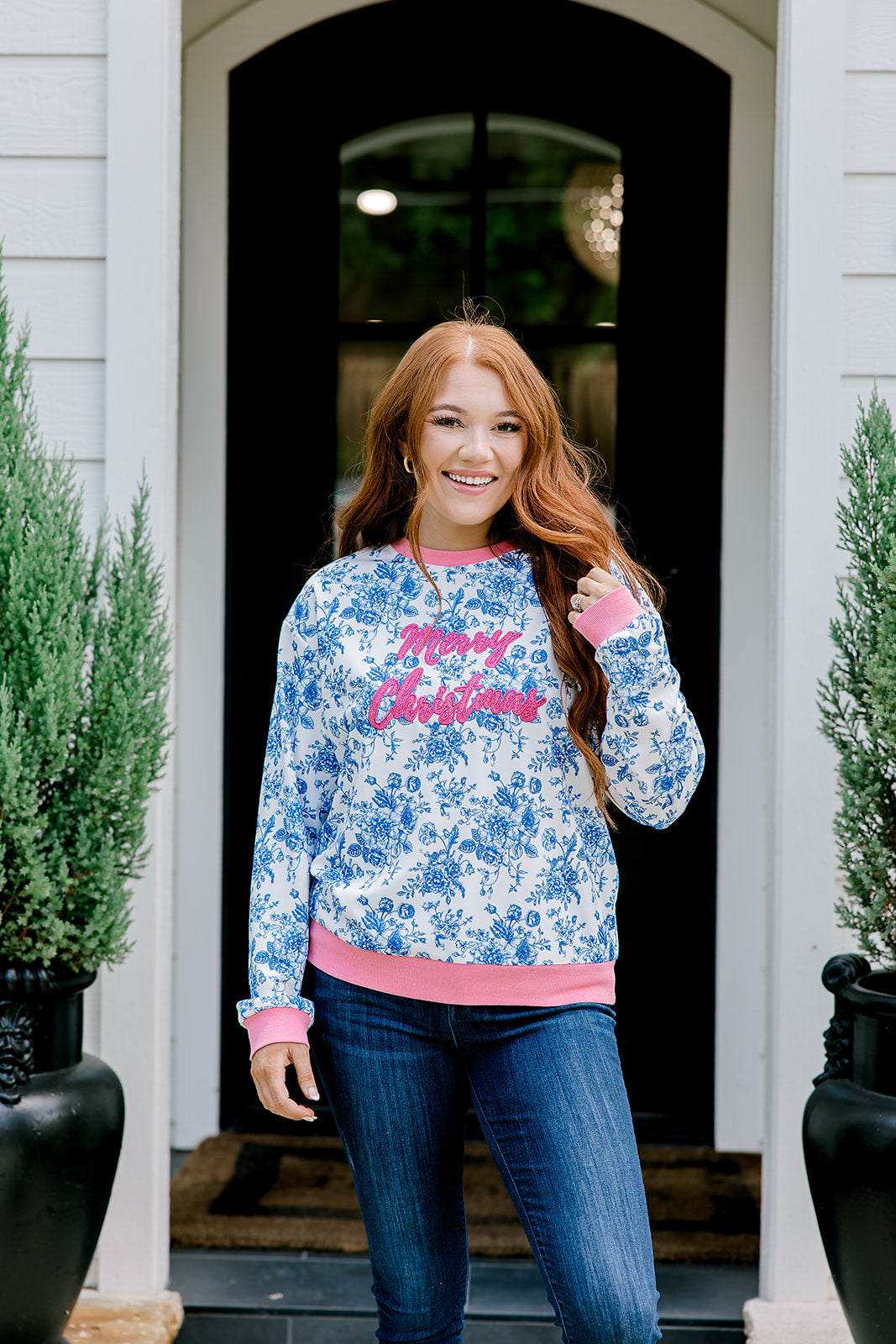 Woman with red hair wearing a blue floral sweatshirt with pink accents and "Merry Christmas" embroidered on front. Model paired this top with denim jeans. 