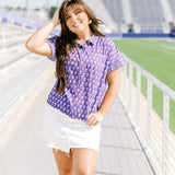 Woman with dark hair standing in a front profile position wearing the Stevie Top at a football field. The focal point of this image is the front graphics of the top as well as the ruffle striped cuff. The model paired this top with a white denim skirt. 