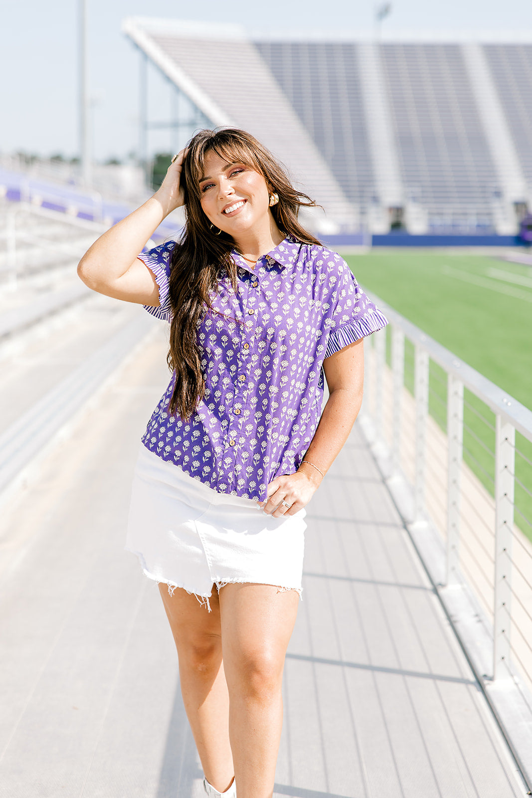 Woman with dark hair standing in a front profile position wearing the Stevie Top at a football field. The focal point of this image is the front graphics of the top as well as the ruffle striped cuff. The model paired this top with a white denim skirt. 