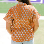 Woman with dark hair facing backwards wearing a short-sleeve, burnt orange top with flower graphics and light-wash denim shorts. 